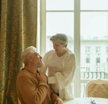 Two people by a bright window, one standing beside a seated person in a cozy room.