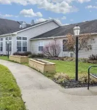 Courtyard walkway beside a white building with benches, lamp posts, and flower beds under a partly cloudy sky