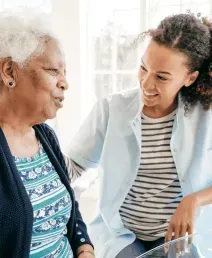 Two women talking in a bright kitchen, one seated and smiling while the other leans in beside her.