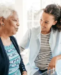 Two women talking in a bright kitchen, one seated and smiling while the other leans in beside her.