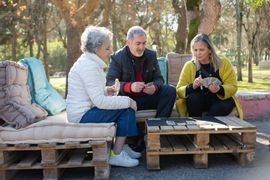 Three people chatting on pallet benches outdoors, holding cups in a sunny park.