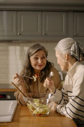 Two women chatting in a kitchen while tossing salad in a glass bowl