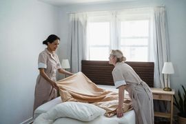 Two women making a bed in a bright bedroom, pulling a beige blanket into place.