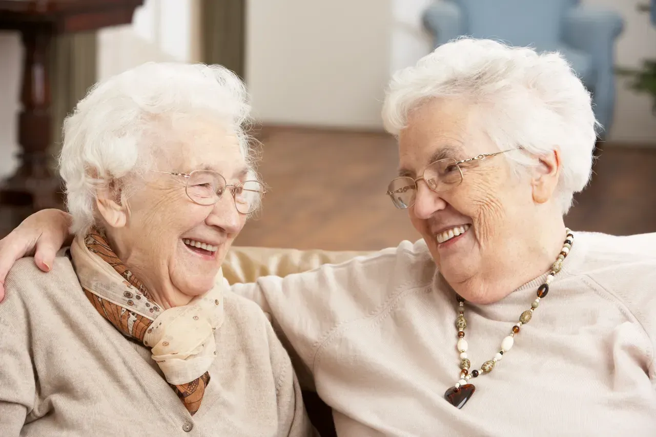 Two smiling seniors sitting close together and embracing indoors, wearing light sweaters and glasses