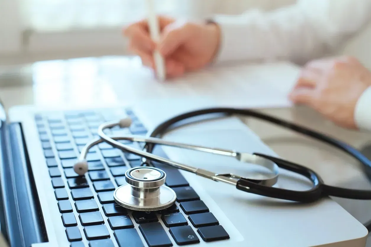 Stethoscope on laptop keyboard with a doctor’s hand writing in the background