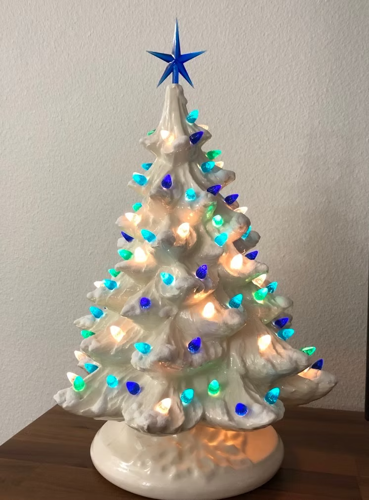 Ceramic Christmas tree with blue star topper, lit with green, blue, and white lights, on a white base.