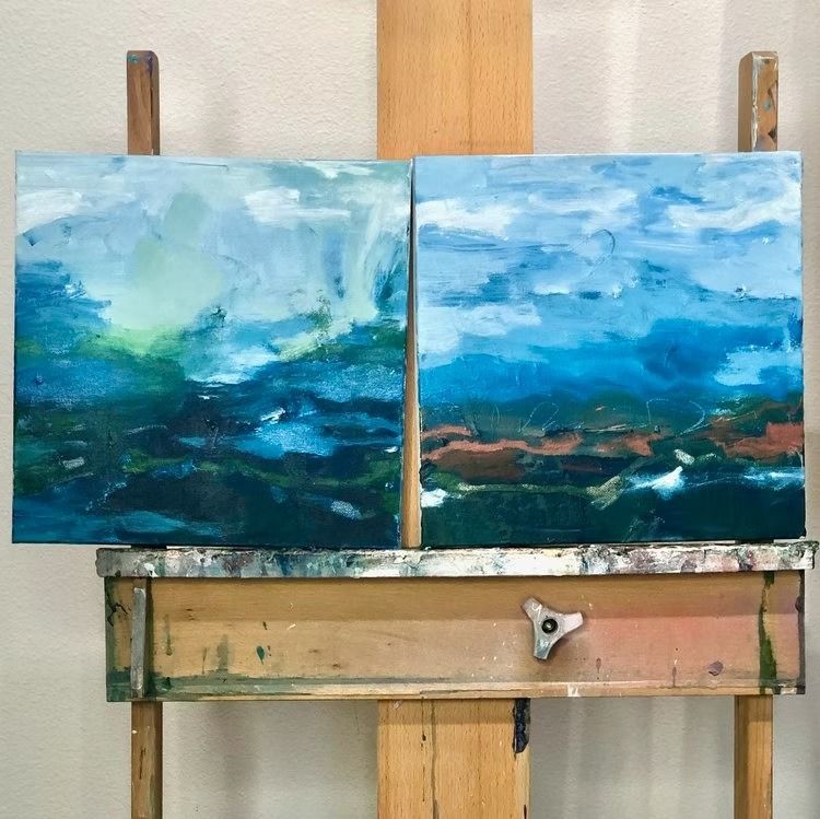 Two abstract landscape paintings in shades of blue and green on an easel.