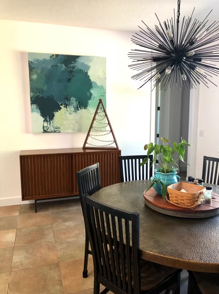 Dining room with dark round table, wooden chairs, and abstract blue-green painting above wooden cabinet.