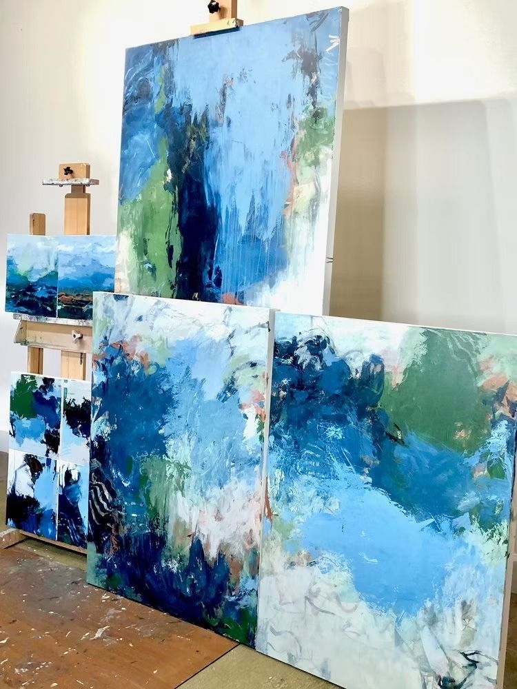 Abstract paintings in shades of blue and green, displayed in a studio setting on easels and the floor.