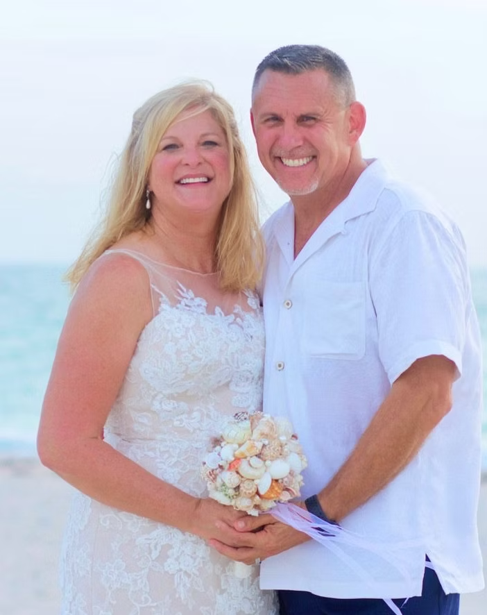 Couple on a beach; woman in white lace dress, holding seashell bouquet, man in white shirt smiling.