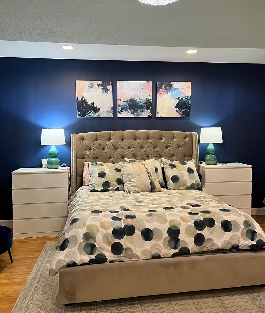 Bedroom with a navy blue wall, tufted headboard, art, and patterned bedding. Two white nightstands flank the bed with green lamps.
