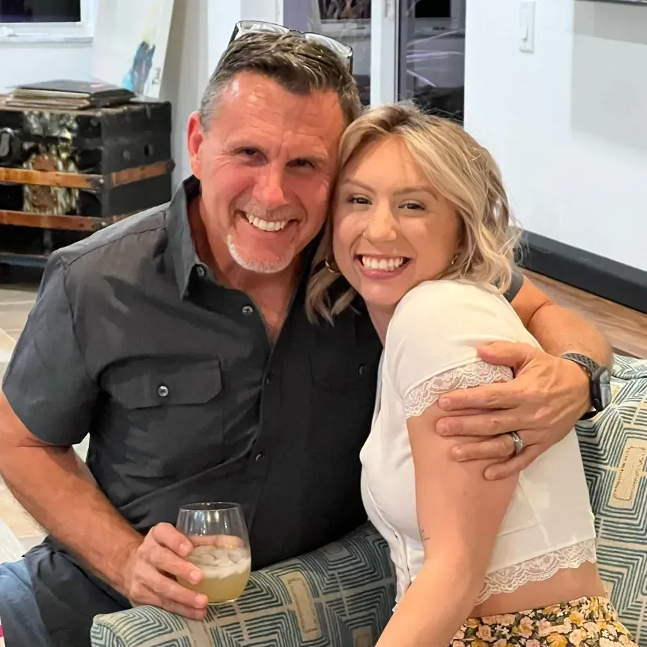 Man hugs a woman; both smile. Man holds a drink. They sit indoors near a couch.