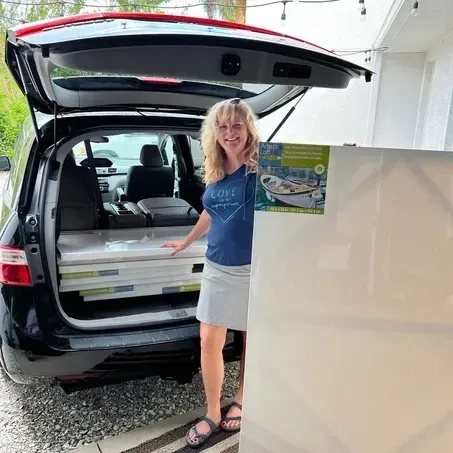 Woman with a blue shirt and gray skirt stands by a car with its trunk open, filled with white rectangular items.