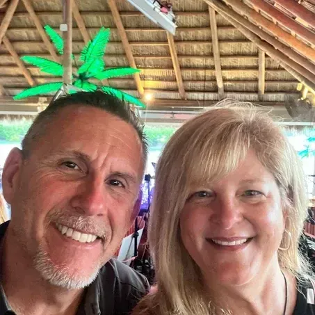 Two smiling people, selfie-style, in tropical-themed setting. Man with short hair, woman with blonde hair.