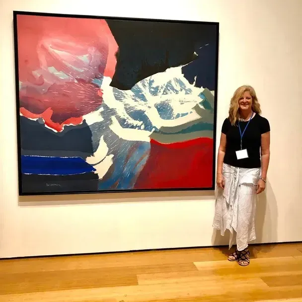 Woman stands next to a large abstract painting with red, blue, and white brushstrokes in a museum setting.