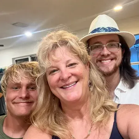 Woman smiles between two men, one with a hat. All are indoors, possibly at a gathering.