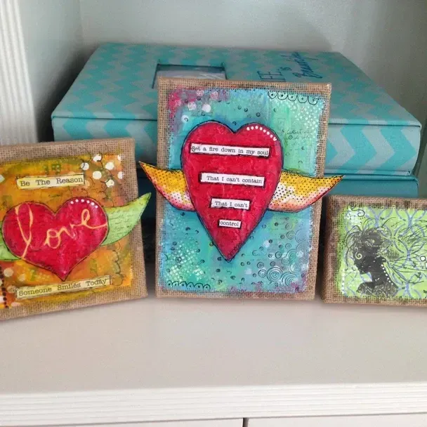 Three mixed-media art pieces with hearts and wings, vibrant colors on a shelf. A blue patterned box in the background.