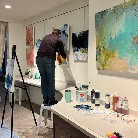 Man adjusting artwork on a wall in a studio with paint supplies and an easel.
