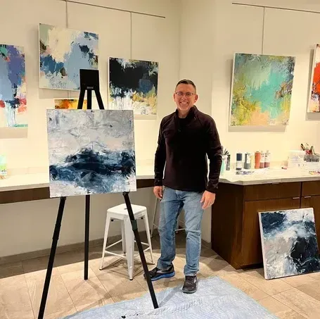 Man in art studio poses beside canvas on easel, surrounded by abstract paintings.