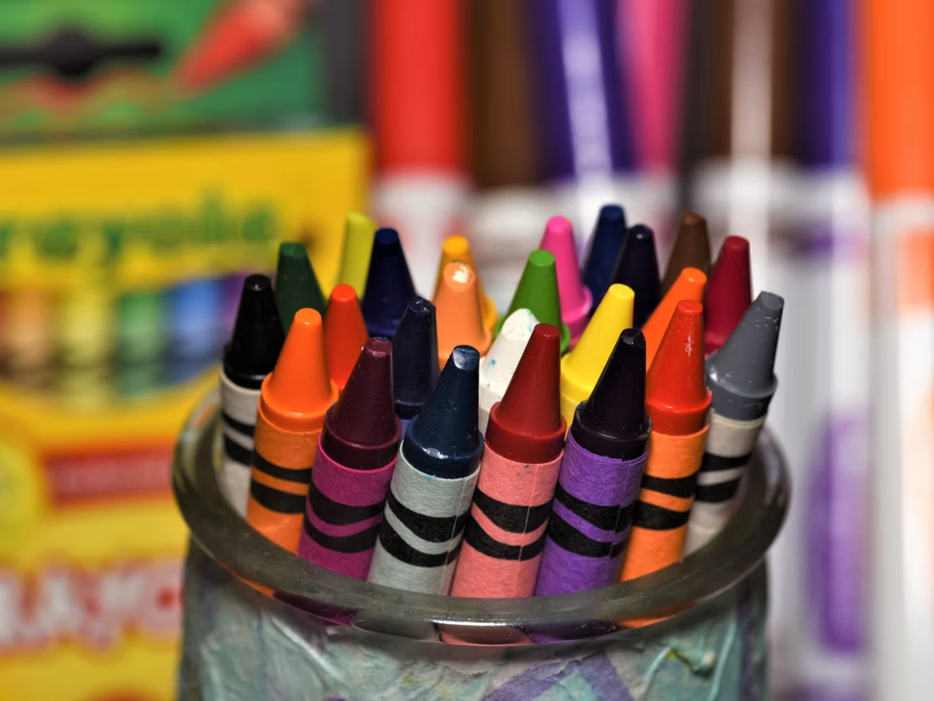 Crayons in a glass jar, various colors, with markers blurred in the background.
