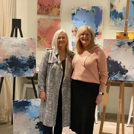 Two women pose in front of abstract paintings; one in a snakeskin coat, the other in a pink sweater and black skirt.