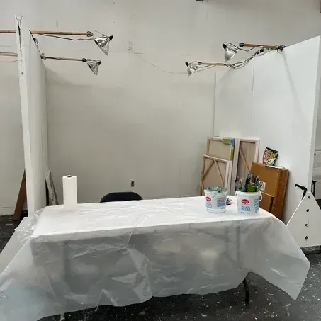 White art studio setup with a table covered in plastic. Paint supplies, art, and lights are visible.