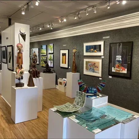 Art gallery interior with paintings, sculptures, and glass art on display under track lighting.