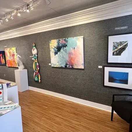 Art gallery interior with framed artwork and sculptures on gray textured walls; hardwood floors.