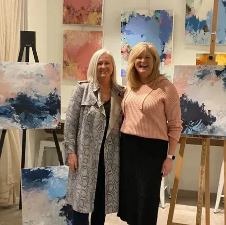 Two women stand by abstract paintings in a gallery. One wears a snake-print coat.