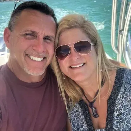 Smiling couple on a boat near water. Man in maroon shirt, woman in sunglasses and patterned top.