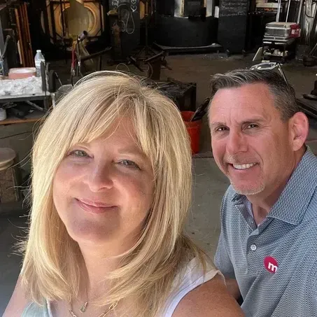 Blond woman and man smile for a selfie in workshop, man wears a polo shirt.