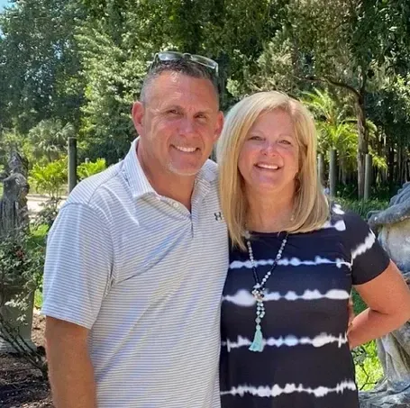 Couple smiling outdoors, man in striped polo shirt, woman in tie-dye dress, trees in background.