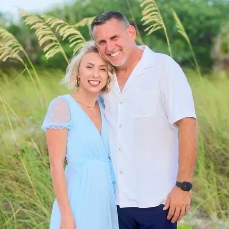 Couple smiling, posing close together outdoors. Woman in light blue dress, man in white shirt, greenery background.