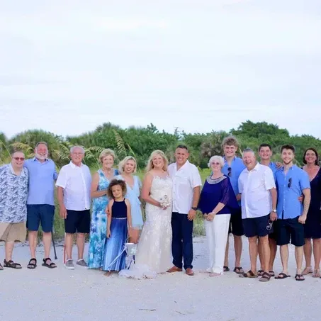 Wedding group on a beach; couple in center, others in blue and white attire, trees and sky background.