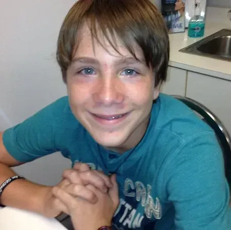 Boy smiling, hands clasped, wearing a teal t-shirt. Kitchen setting, sink in background.