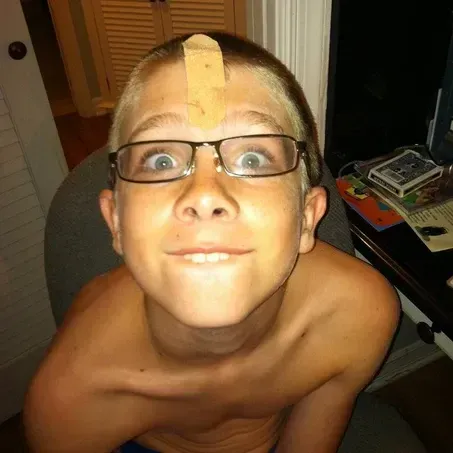 Boy with glasses and band-aid on forehead, making a funny face, indoors.