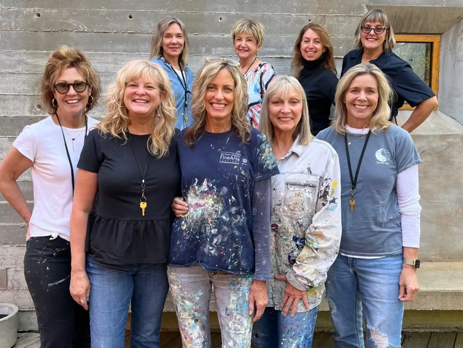 Nine women pose outdoors, in front of concrete steps. Most wear paint-splattered clothing, smiling.