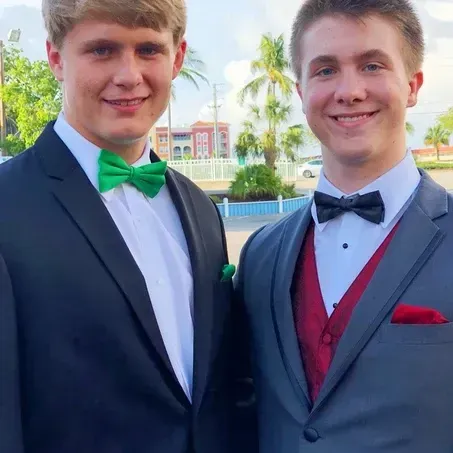 Two young men in formal suits posing outside; one with a green bow tie, the other with a black bow tie and red vest.
