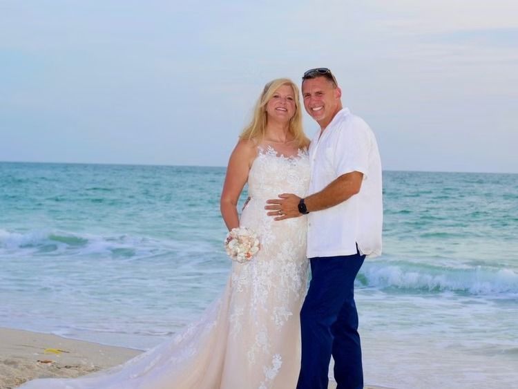 Couple on a beach: Woman in wedding dress, man in white shirt, smiling, posing near ocean waves.