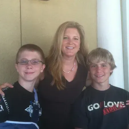 Woman with two boys, all smiling. The woman has long blonde hair and is wearing a dark top. The boys are wearing black t-shirts.