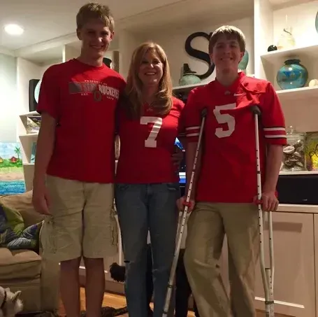 Family of three wearing red Ohio State gear, posed indoors. One son on crutches.