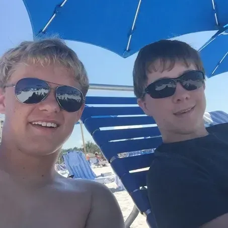 Two people wearing sunglasses sitting on beach chairs under a blue umbrella.