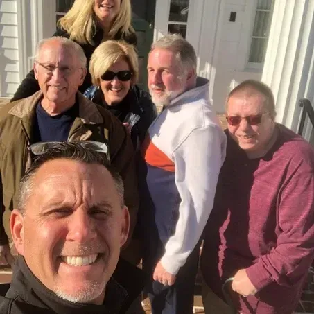 Group of people smiling for a photo outside a white building. Sunlit, with varied clothing.