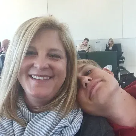 Woman smiles, headshot with blond hair, scarf; man rests on her shoulder, airport setting.