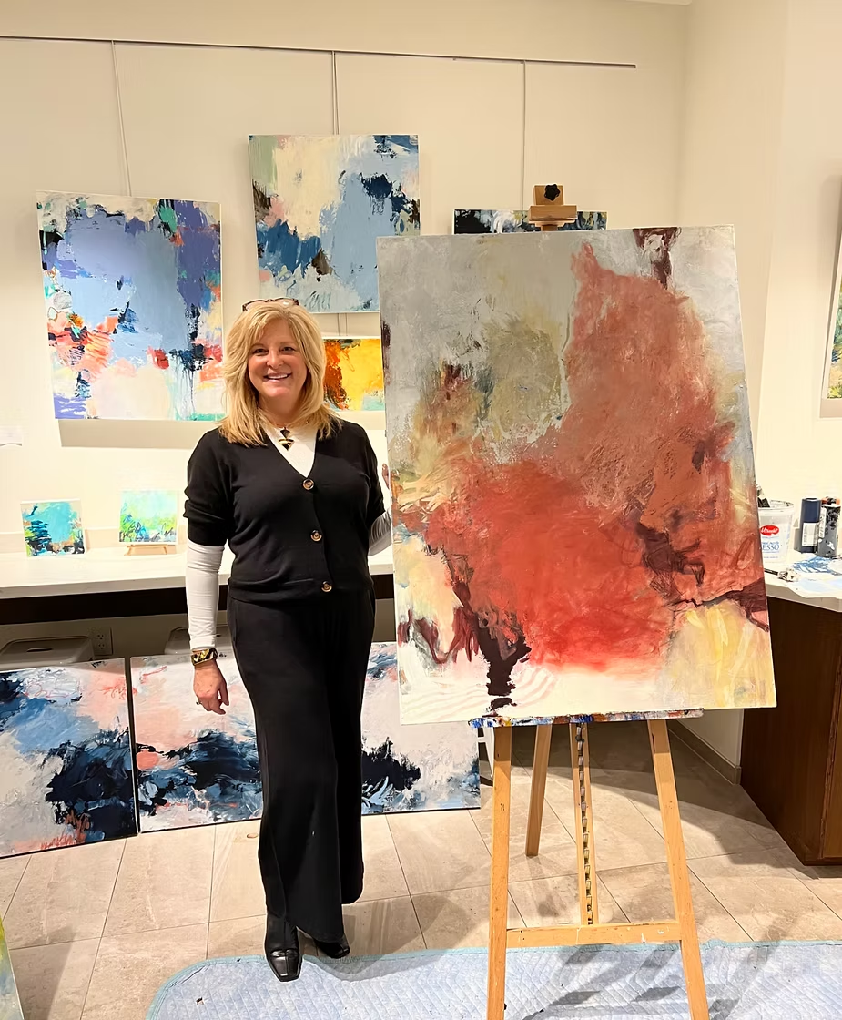 Woman in gallery poses with abstract painting on easel, surrounded by other artwork.