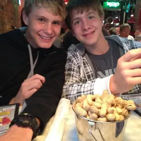 Two young men smiling at a table with a bucket of peanuts in a restaurant.