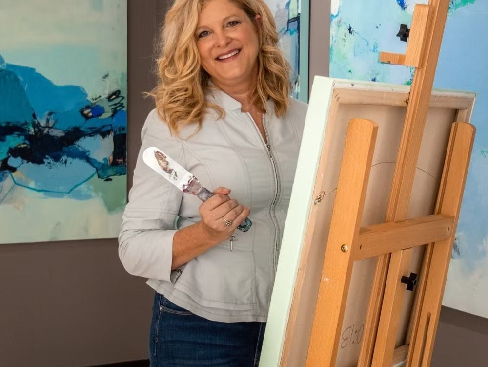 Woman holding a palette knife smiles, standing by canvas on easel with blue paintings in background.