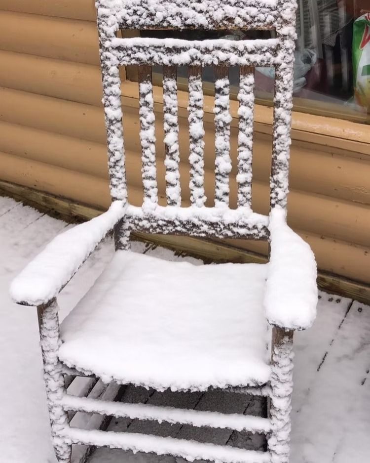 Snow-covered wooden rocking chair on a porch, near a log cabin wall.