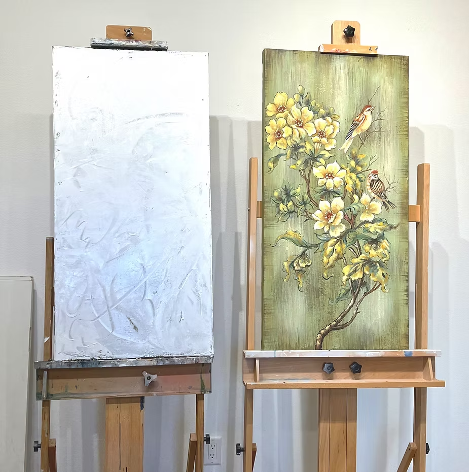 Two canvases on easels: one blank, the other a painting of yellow flowers and birds on a green background.