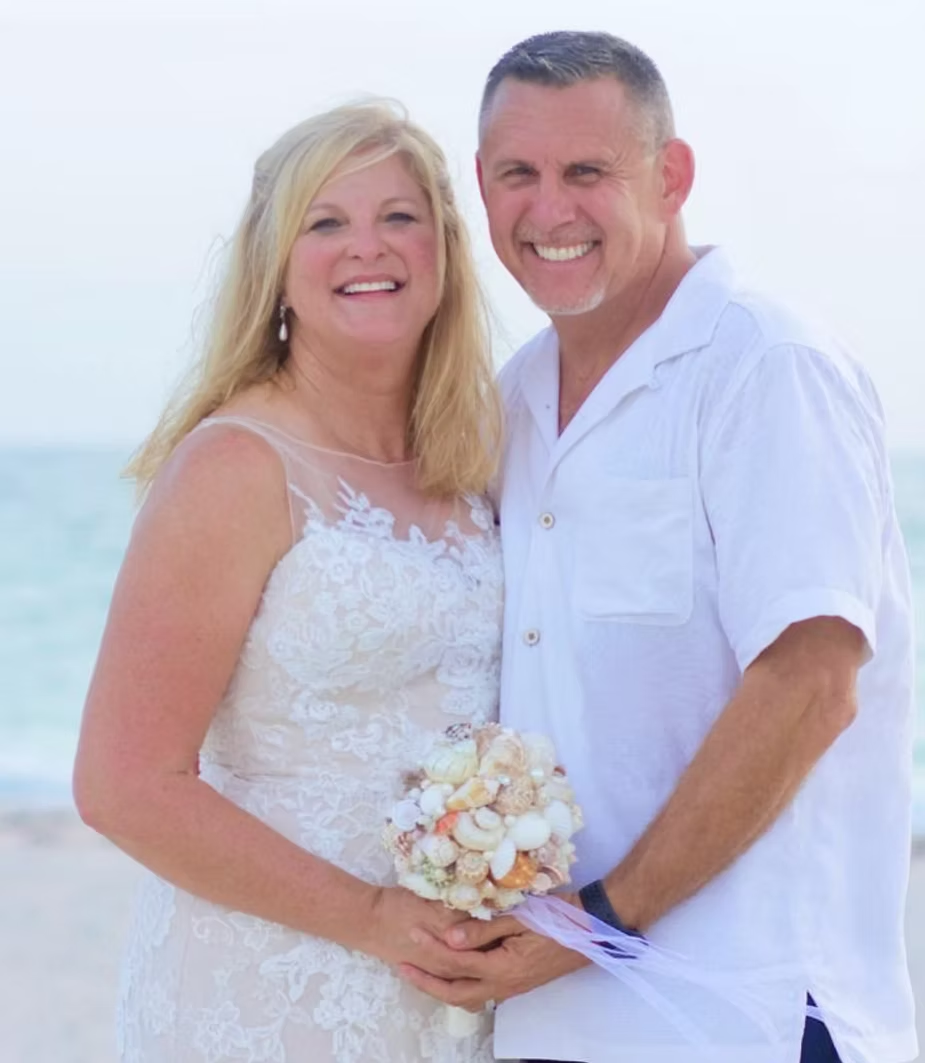 Couple smiling, posing together on a beach, woman in white wedding dress, man in white shirt.